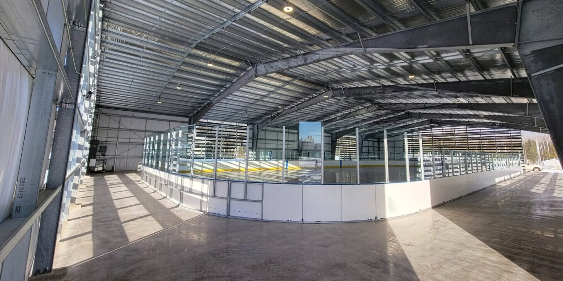 View of an ice rink in the midst of construction, showing large exposed steel beams and insulation. Taken from one of the corners of the building, the outside of the rink boards and glass can be seen in the foreground.